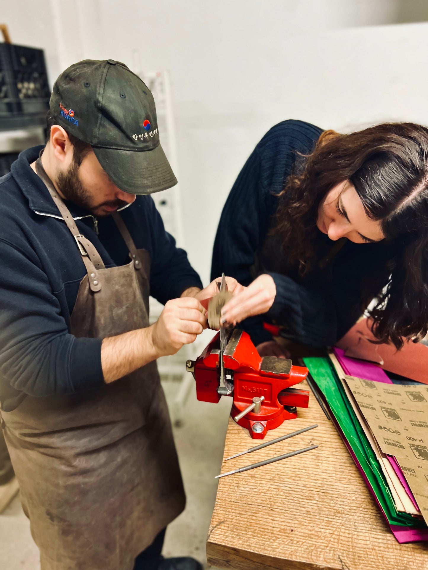 Couple working together to finish a hand-forged knife during beginner blacksmithing workshop at Oldboy Metal Co. in Toronto, sanding blade at vise station.