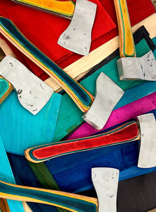 A collection of custom hand-forged axe heads paired with vibrant, multicolored wooden handles, laid out on matching dyed wood panels at Oldboy Metal Co. 