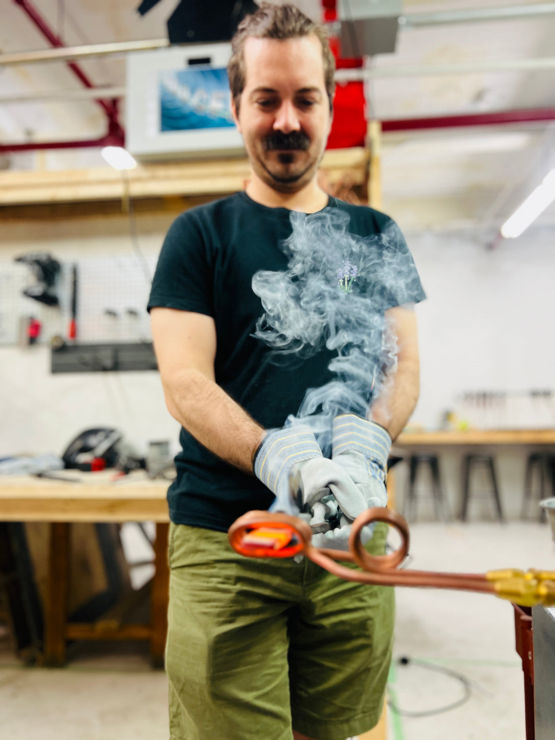Man holding a glowing red-hot piece of steel with tongs while using an induction forge, as smoke rises dramatically in the blacksmithing workshop at Oldboy Metal Co..