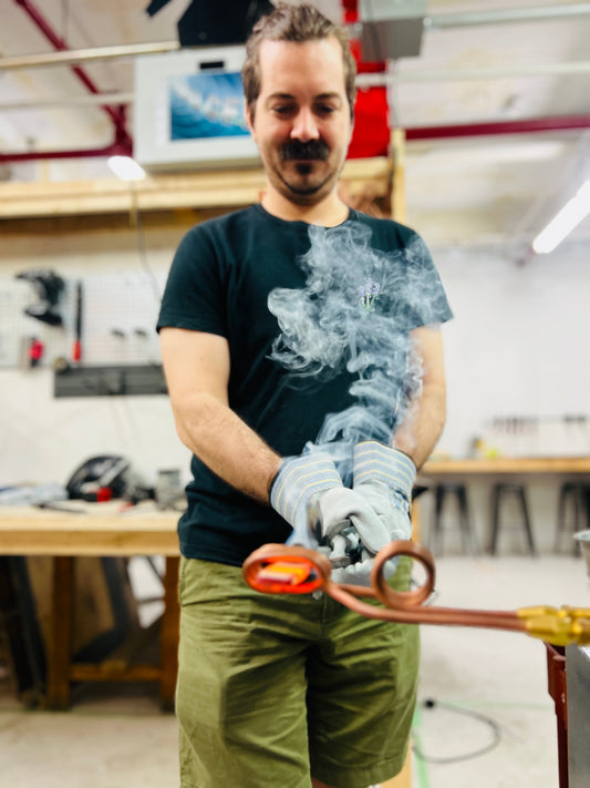 Man holding a glowing red-hot piece of steel with tongs while using an induction forge, as smoke rises dramatically in the blacksmithing workshop at Oldboy Metal Co..