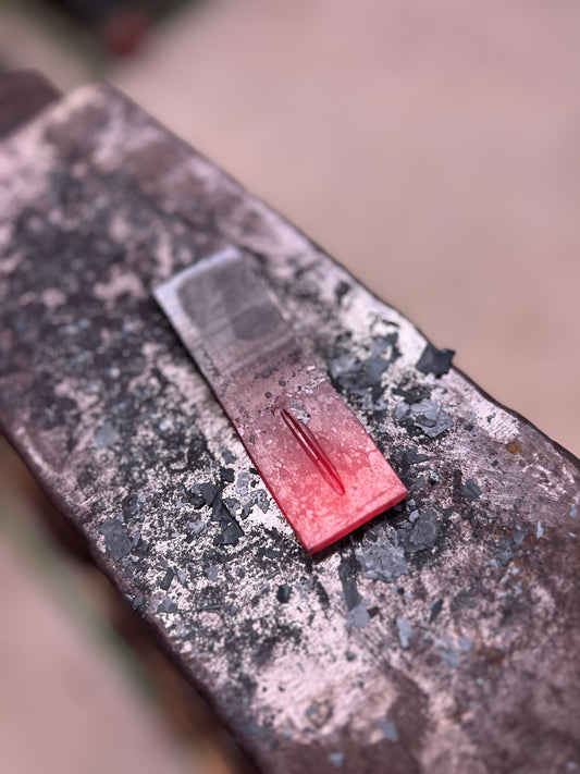 Heated steel flat bar on anvil at Oldboy Metal Co. in Toronto, showing the start of punching the eye for a bottle opener during beginner blacksmithing workshop.