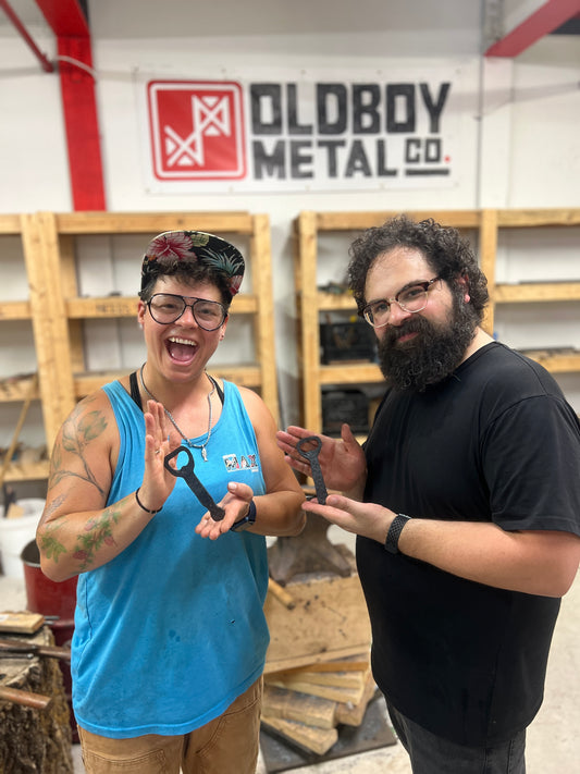 Beginner blacksmithing workshop in Toronto at Oldboy Metal Co. with two participants holding hand-forged bottle openers they made during the class